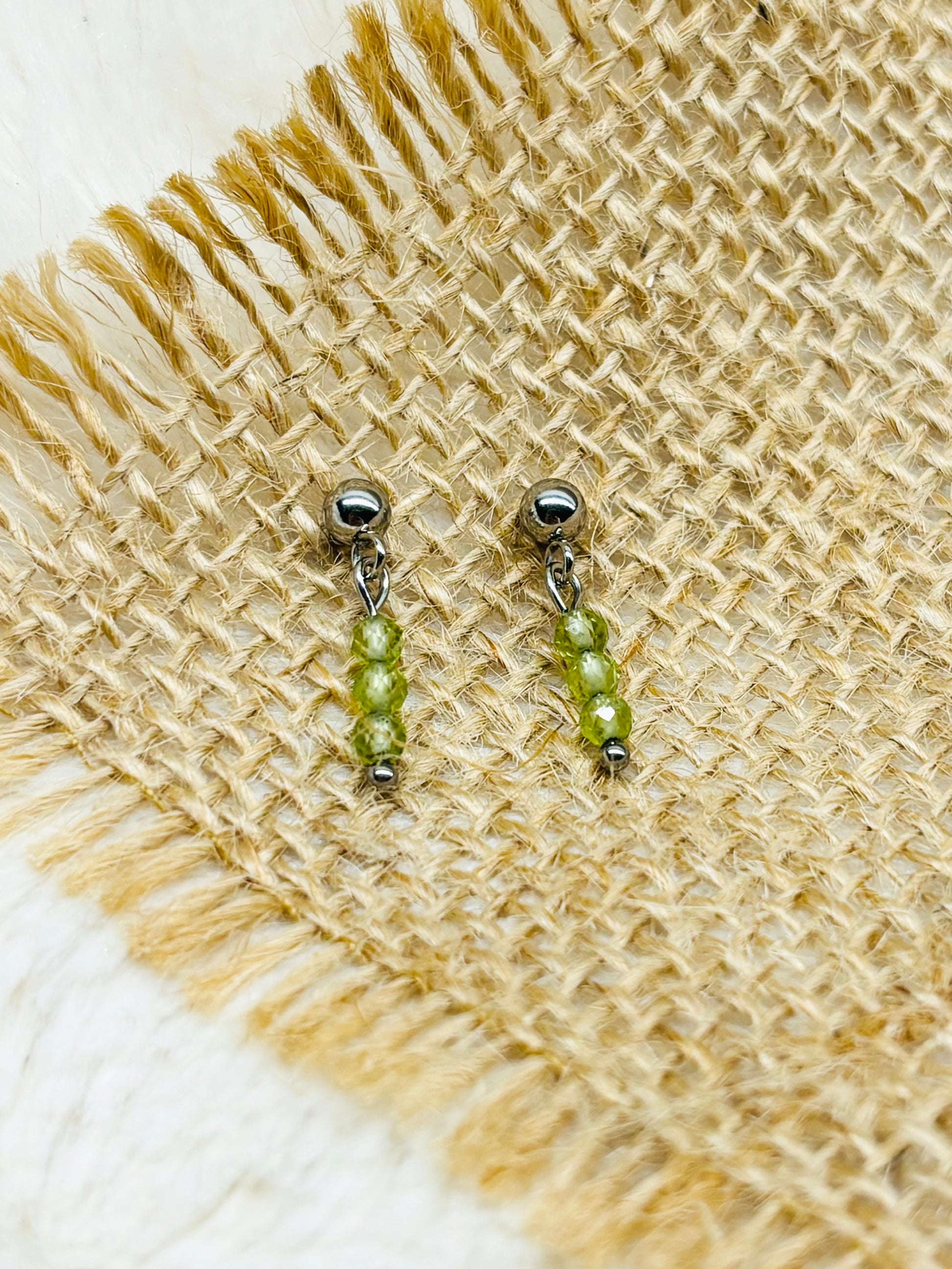 Boucles d'oreille LALY, avec pierres de Péridot, exposées sur un fond en toile de jute.