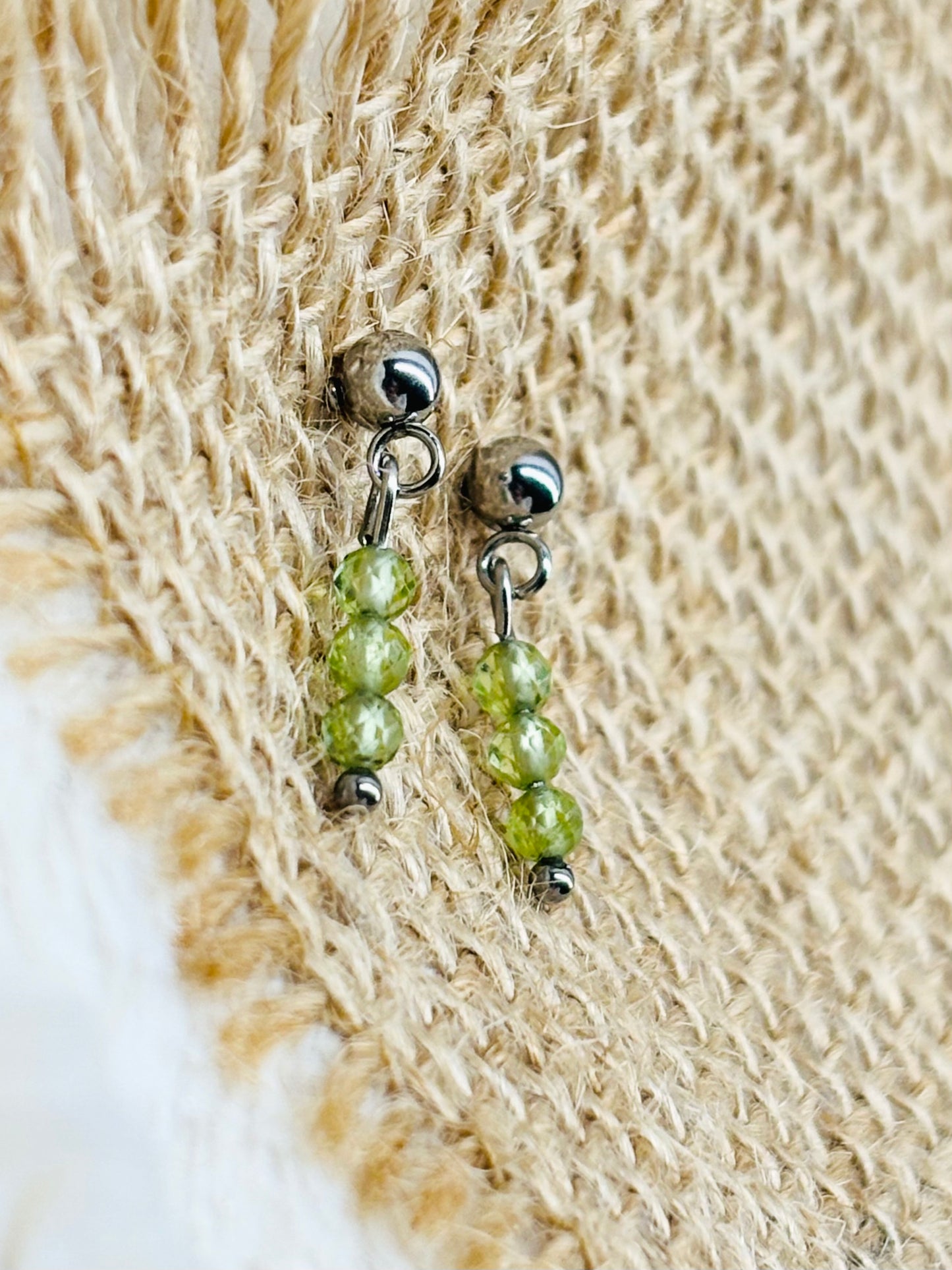 Boucles d'oreille LALY, avec pierres de Péridot, exposées sur un fond en toile de jute.