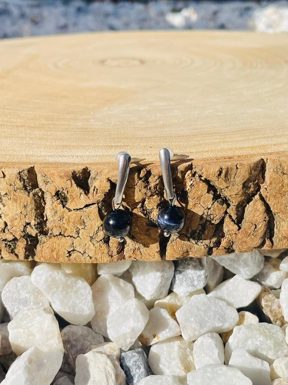 Boucles d'oreille MADELEINE, avec pierre d'Oeil de Faucon, exposées sur un rondin de bois.