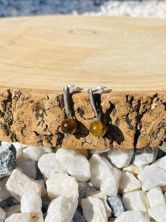 Boucles d'oreille MADELEINE, avec pierre d'Oeil de Tigre, exposées sur un rondin de bois.