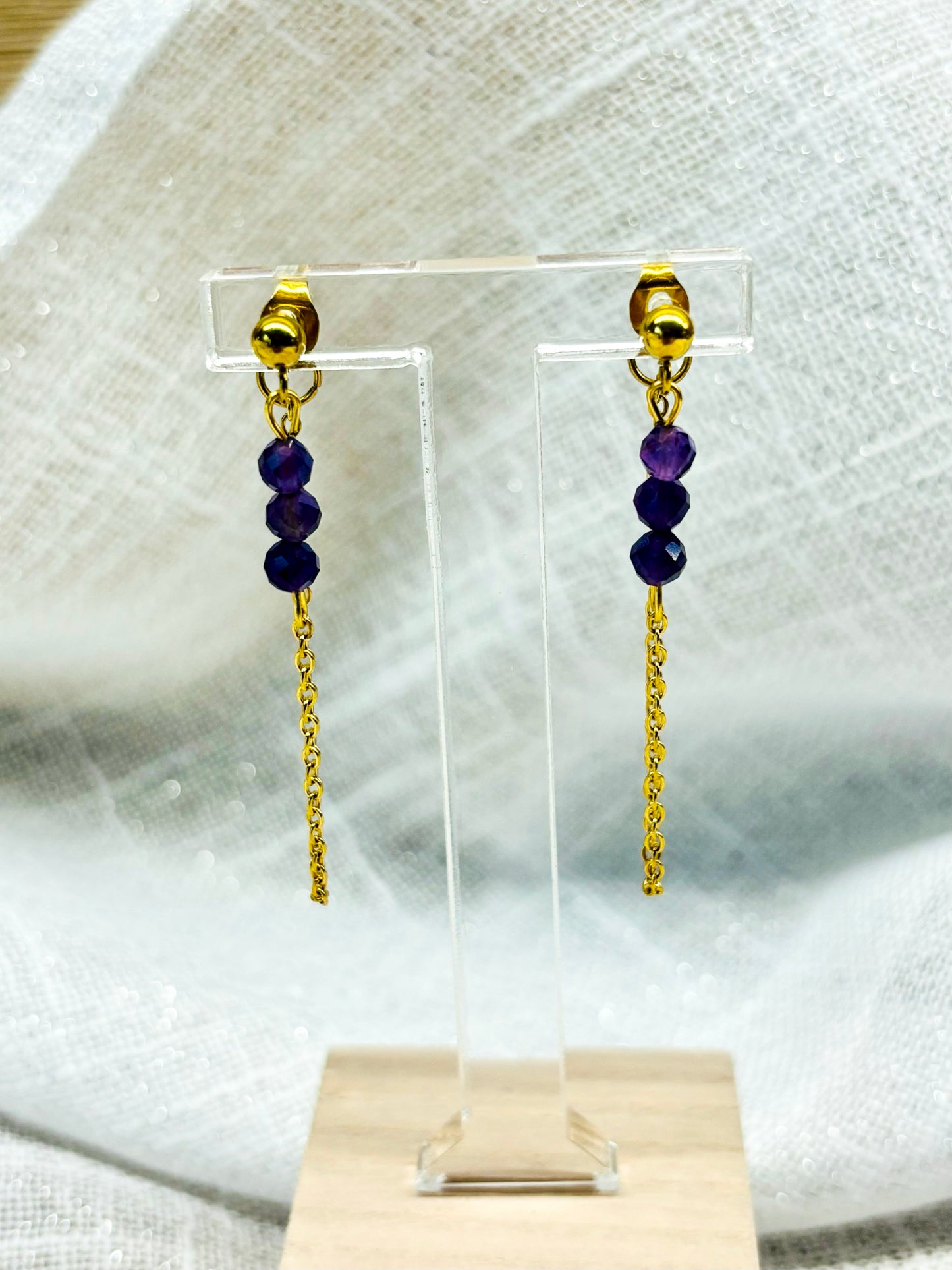 Boucles d'oreille MALIA, de couleur dorée avec pierres d'Améthyste, exposées devant un fond en lin.