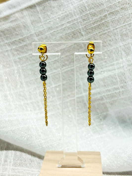 Boucles d'oreille MALIA, de couleur dorée avec pierres d'hématite, exposées sur un fond en lin.