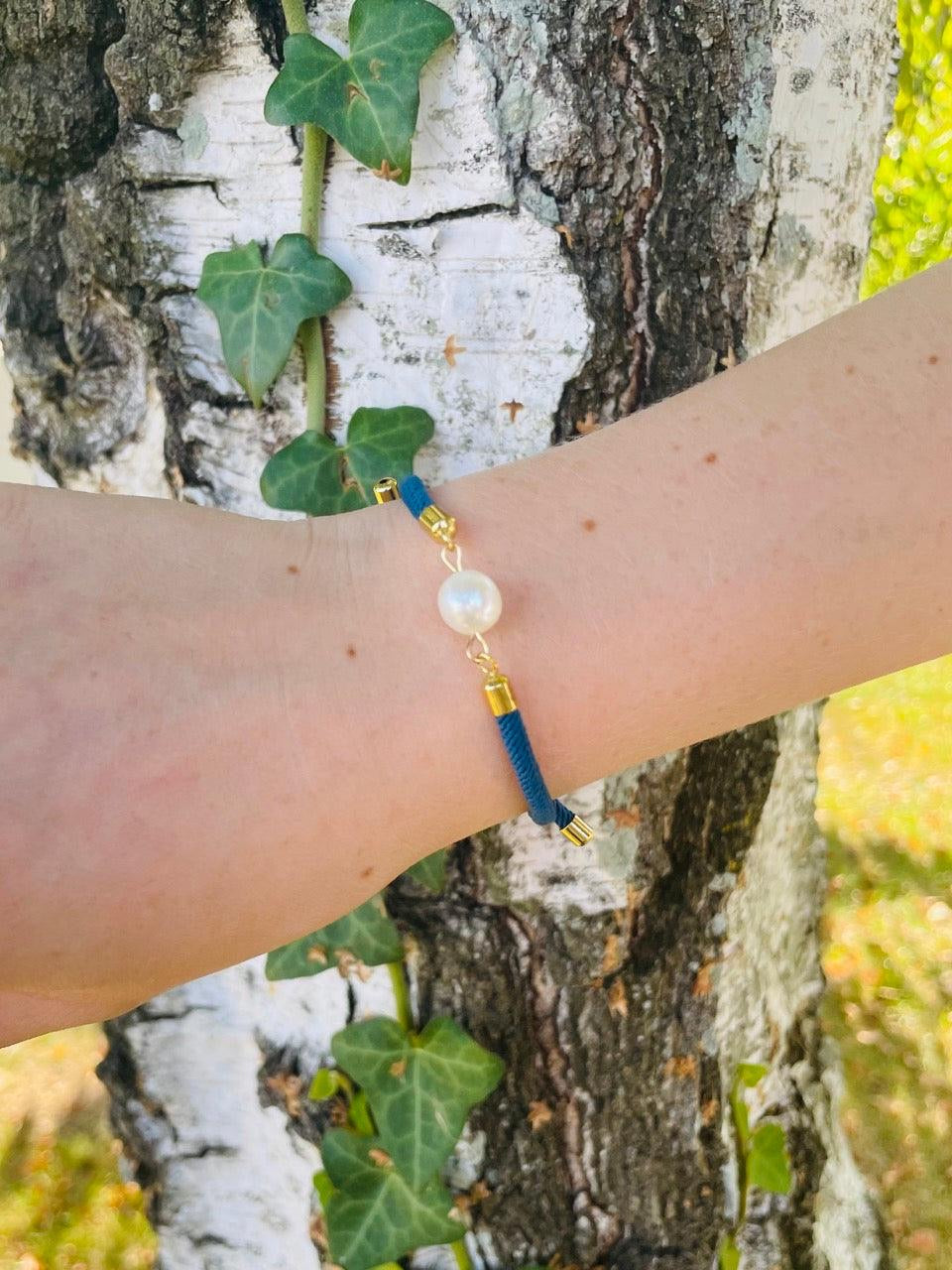 Bracelet MIA, avec cordon de couleur bleu jean et perle de culture, porté par un poignet de femme devant un arbre.