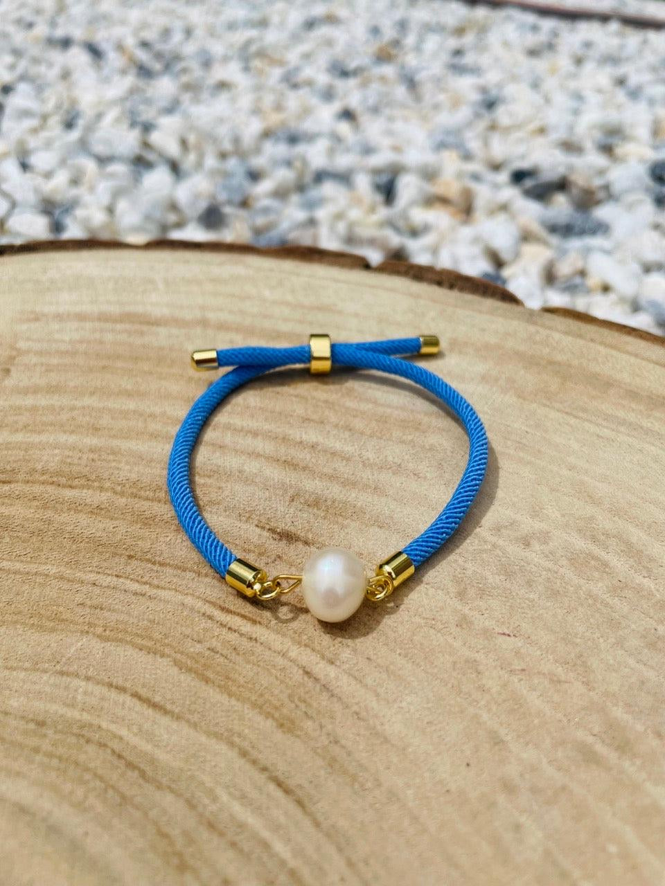 Bracelet MIA avec cordon de couleur bleu et perle de culture, exposé sur un fond en bois et graviers blancs.