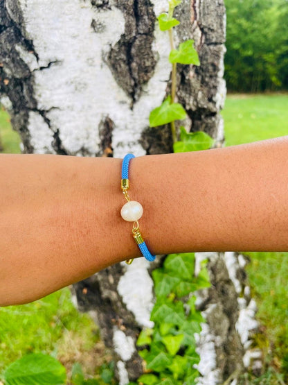 Bracelet MIA avec cordon de couleur bleu et perle de culture, porté sur un poignet de femme devant un arbre.