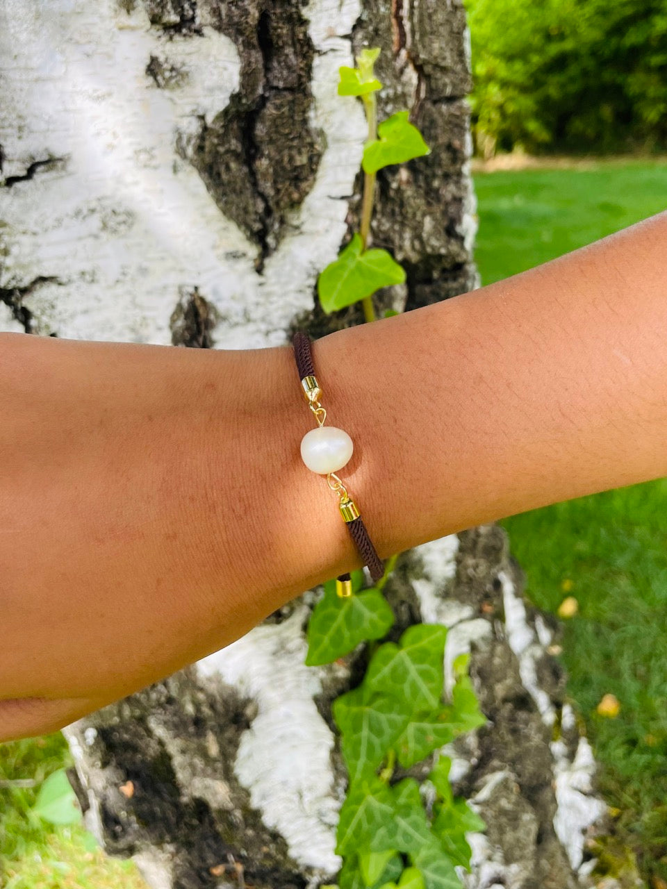 Bracelet MIA en cordon de couleur chocolat et perle de culture, porté sur un poignet de femme devant un arbre