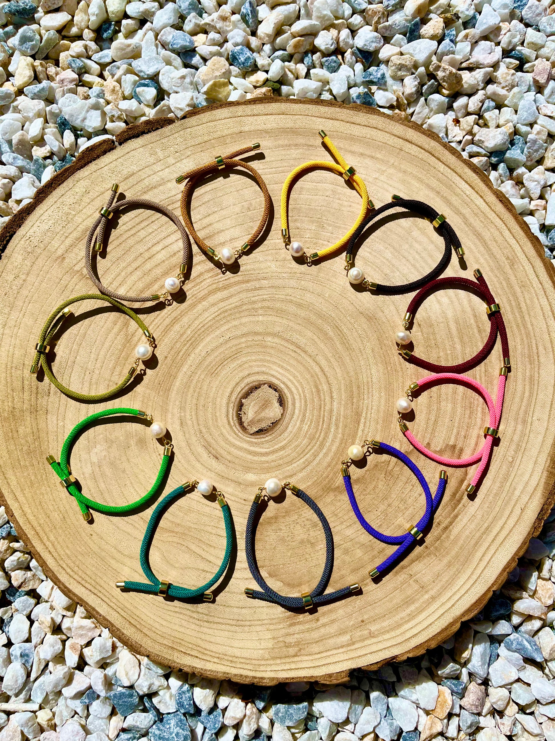 Bracelets MIA, avec cordon de couleur vieux rose et perle de culture, exposés sur une planche en bois et un fond en graviers blancs.