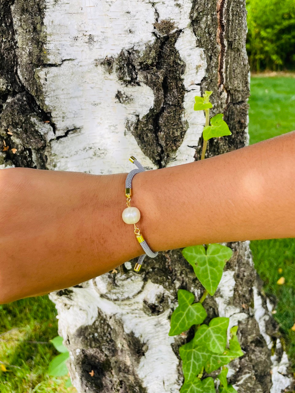 Bracelet MIA, en cordon de couleur gris souris et perle de culture, porté sur un poignet de femme devant un arbre.