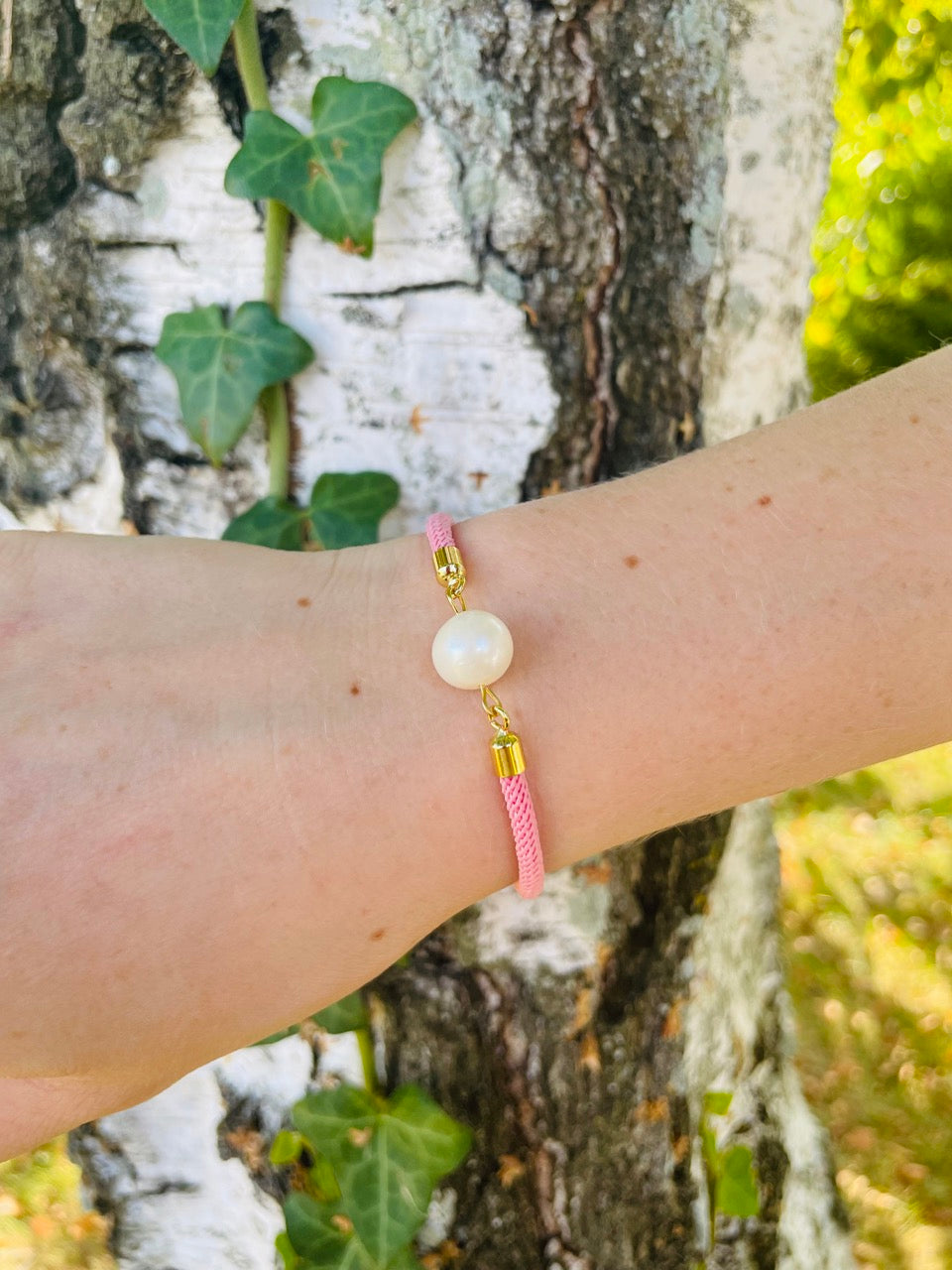 Bracelet MIA, avec cordon de couleur rose clair et perle de culture, porté sur un poignet de femme devant un arbre.