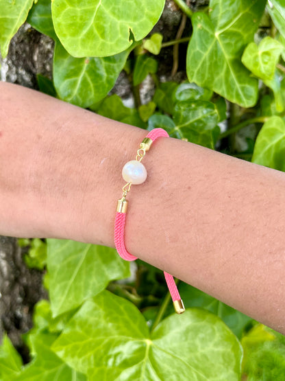 Bracelet MIA avec cordon de couleur rose fluo et perle de culture, porté par un poignet de femme sur des feuilles.