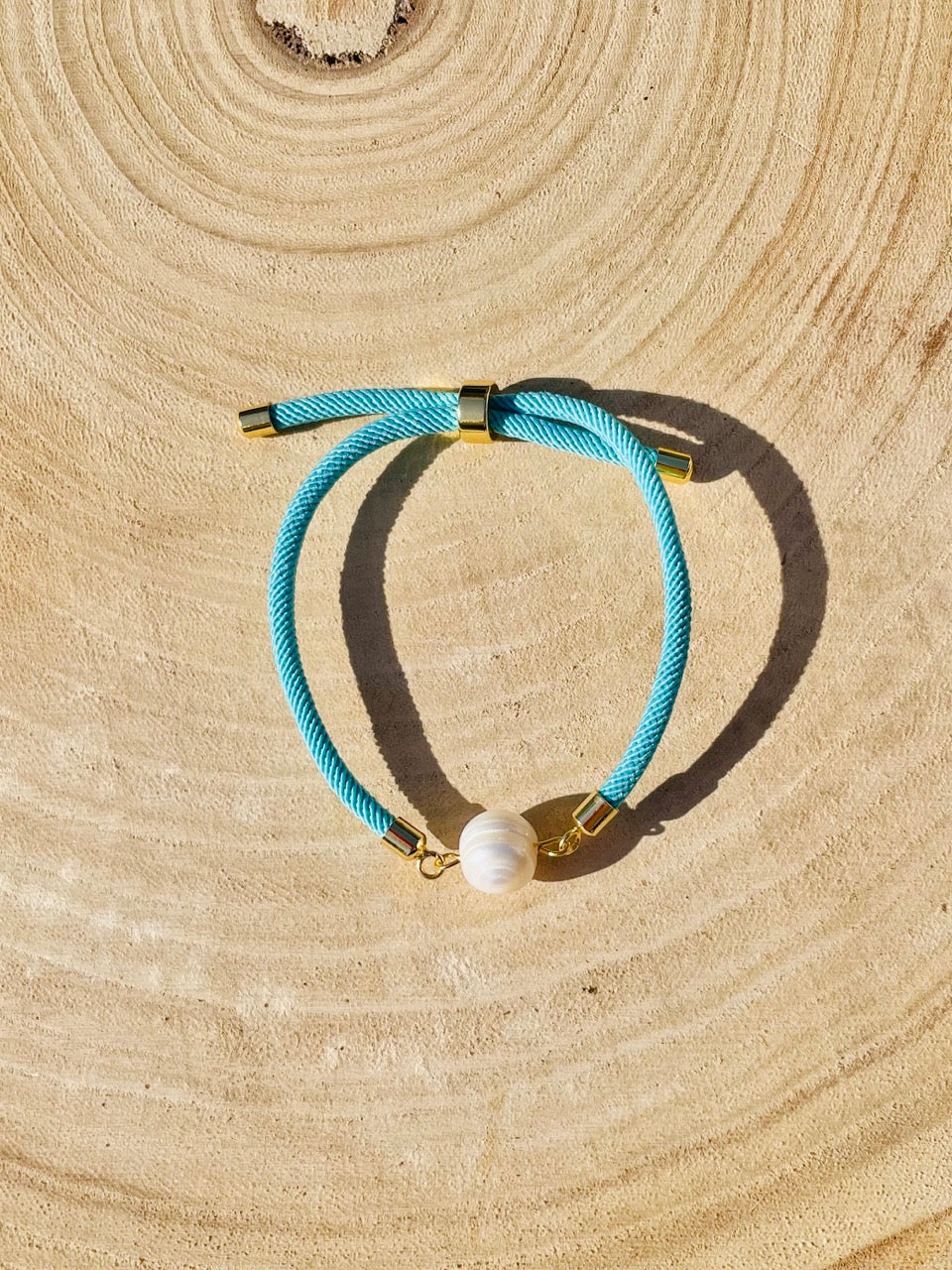 Bracelet MIA, avec cordon de couleur turquoise et perle de culture, exposé sur une planche en bois et un fond de graviers blancs.