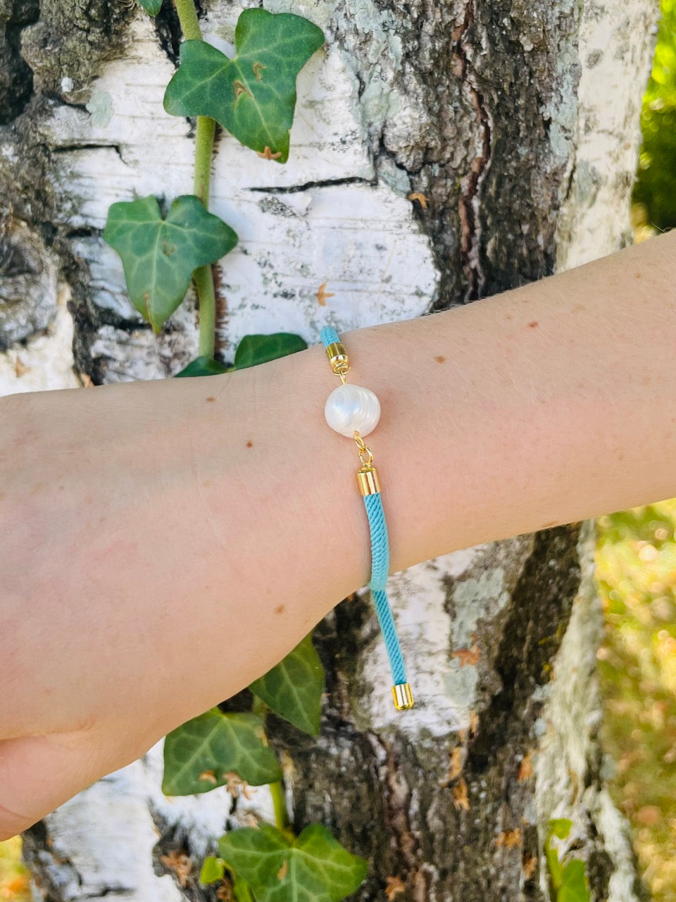 Bracelet MIA, avec cordon de couleur turquoise et perle de culture, porté sur un poignet de femme devant un arbre.