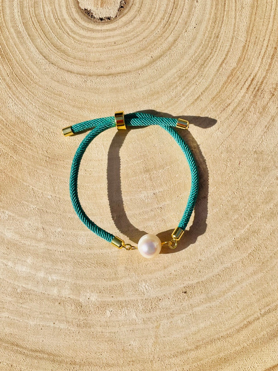 Bracelet MIA, avec cordon de couleur vert émeraude et perle de culture, exposé sur un fond en bois et graviers blancs.