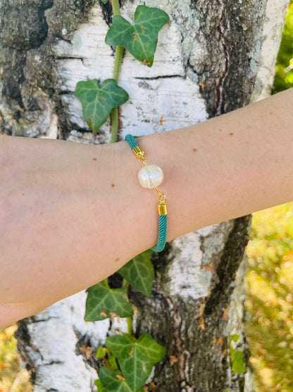 Bracelet MIA, avec cordon de couleur vert émeraude et perle de culture, porté sur un poignet de femme devant un arbre.