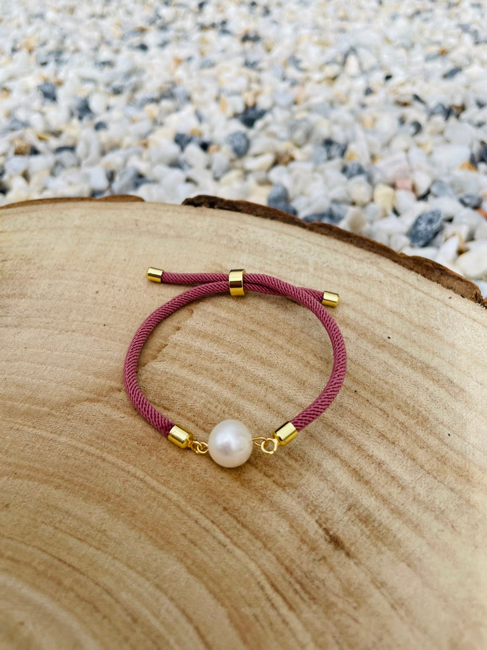 Bracelet MIA, avec cordon de couleur vieux rose et perle de culture, exposé sur une planche en bois et un fond en graviers blancs.