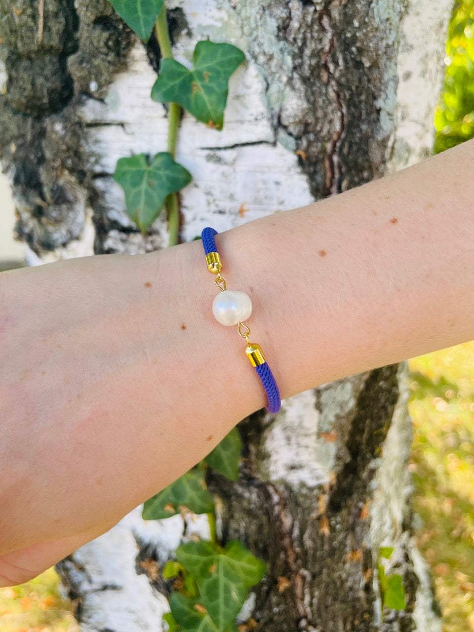 Bracelet MIA, avec cordon de couleur violette et perle de culture, porté sur un poignet de femme devant un arbre.