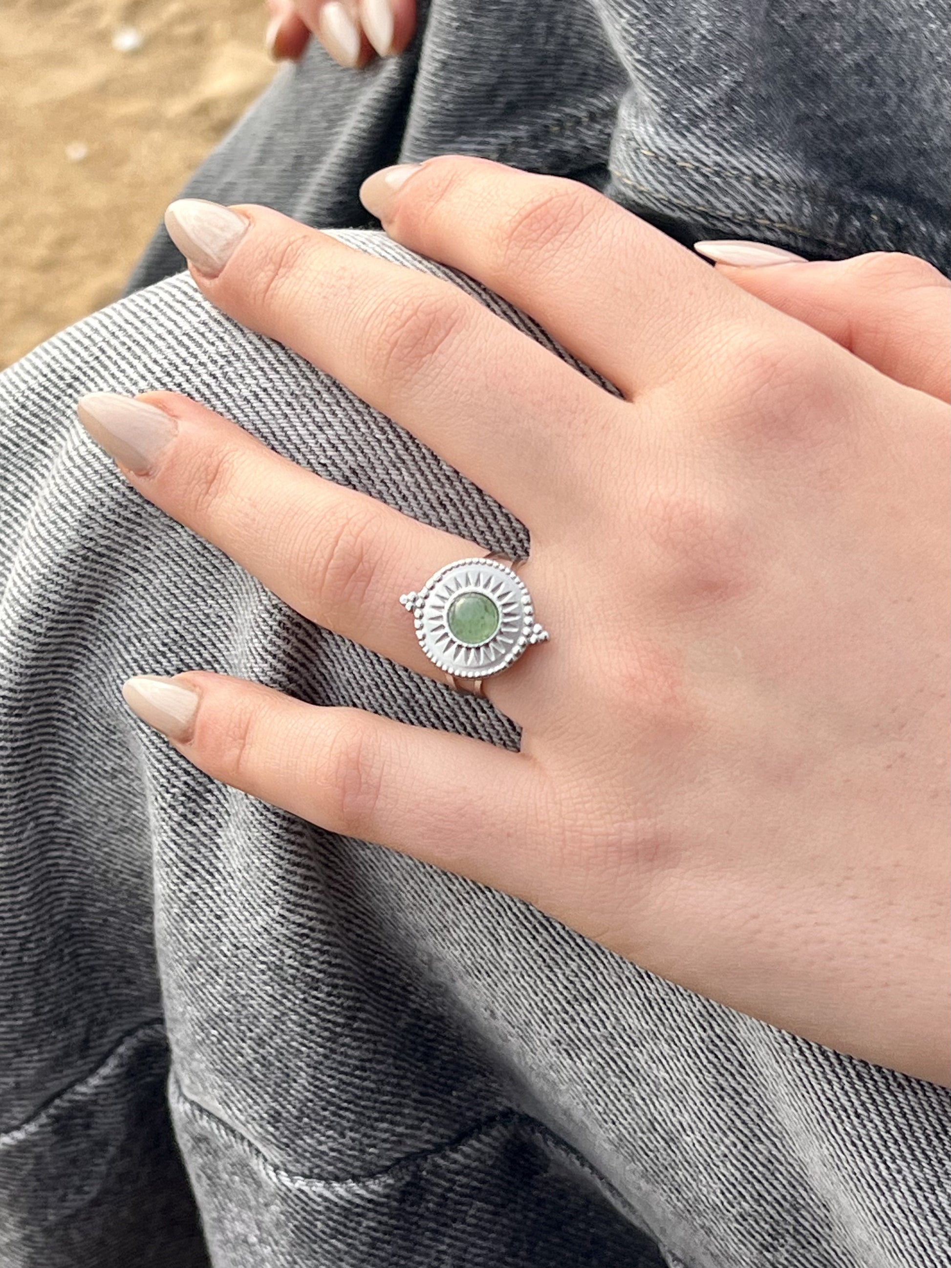 Bague ZÉLIE en acier inoxydable argent, avec pierre d'Aventurine, portée par une main de femme sur la plage.