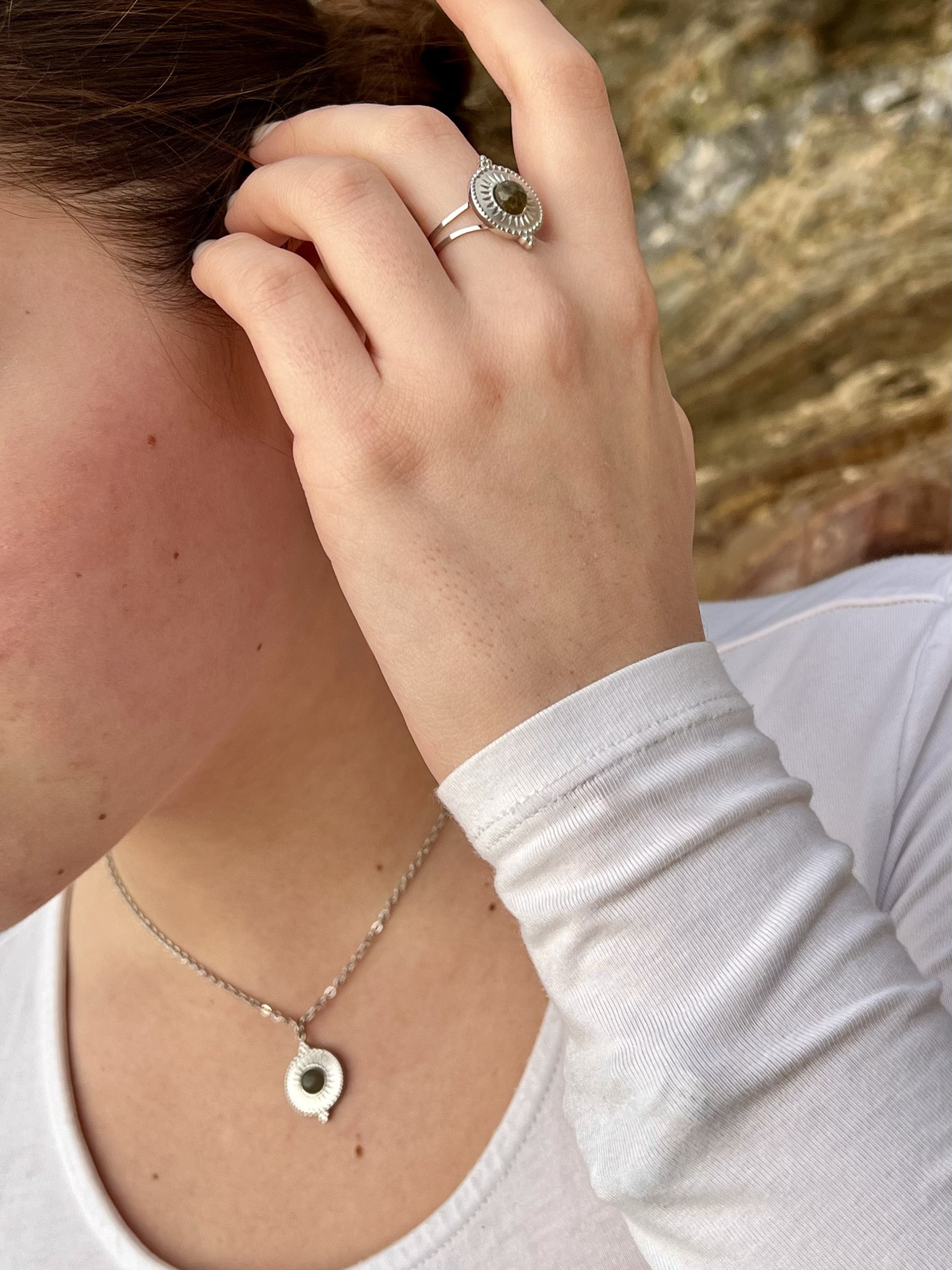 Bague et Collier ZÉLIE en acier inoxydable argent, avec pierre de Labradorite, portés par une femme dans les rochers.