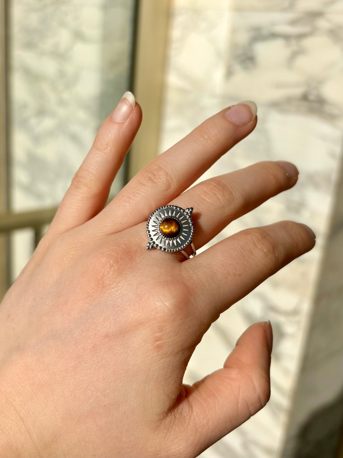 Bague ZÉLIE en acier inoxydable argent avec pierre d'oeil de tigre, porté sur une main de femme avec fond en marbre.