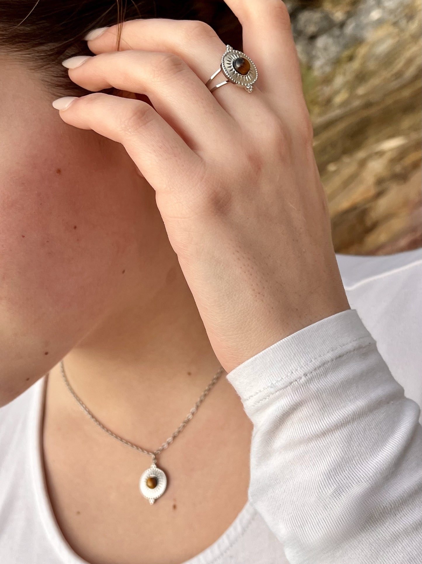 Bague et collier ZÉLIE en acier inoxydable argent avec pierre d'oeil de tigre, porté par une femme dans les rochers.