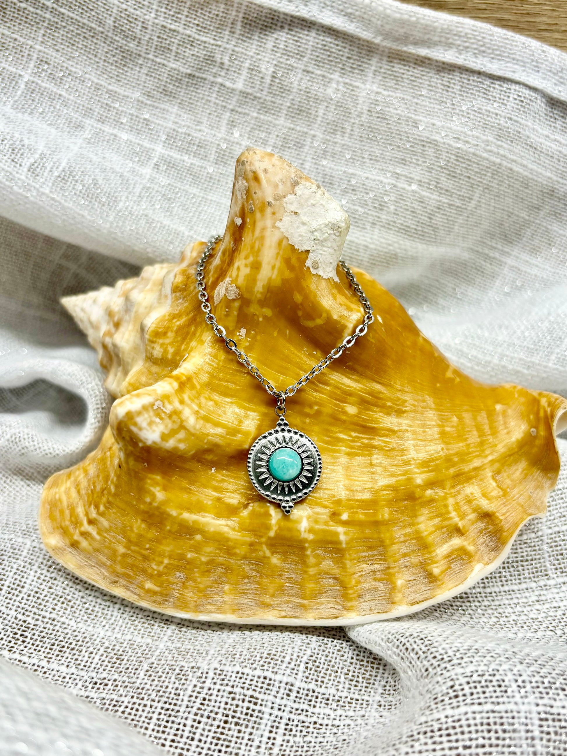 Collier ZÉLIE en acier inoxydable argent, avec pierre d'Amazonite, exposé sur un coquillage et fond en lin blanc.