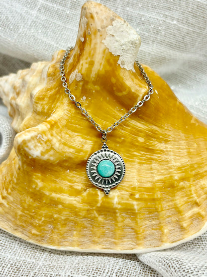 Collier ZÉLIE en acier inoxydable argent, avec pierre d'Amazonite, exposé sur un coquillage et fond en lin blanc.